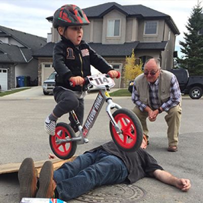 Boy jumps over adult laying on the ground on his Strider 12 Pro while another adult looks on