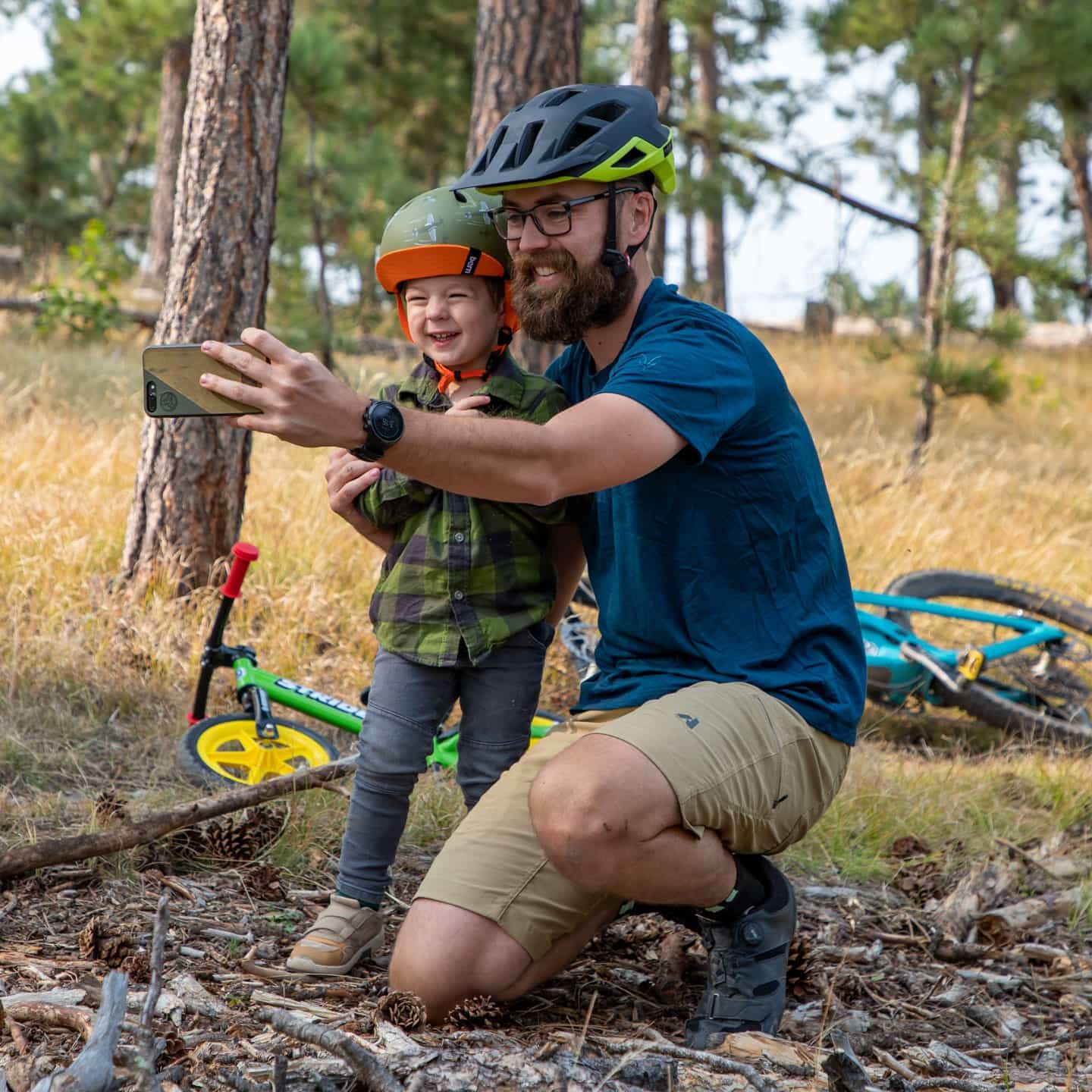 Father takes a selfie with his son in the woods with bikes on the ground in the background