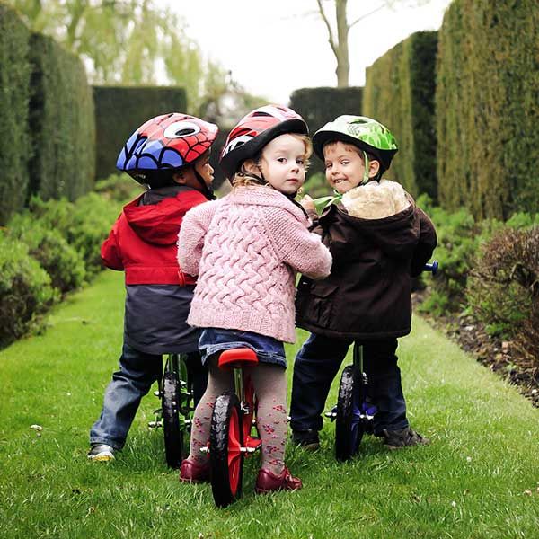 Three kids ride Strider bikes through tall green hedges in a garden