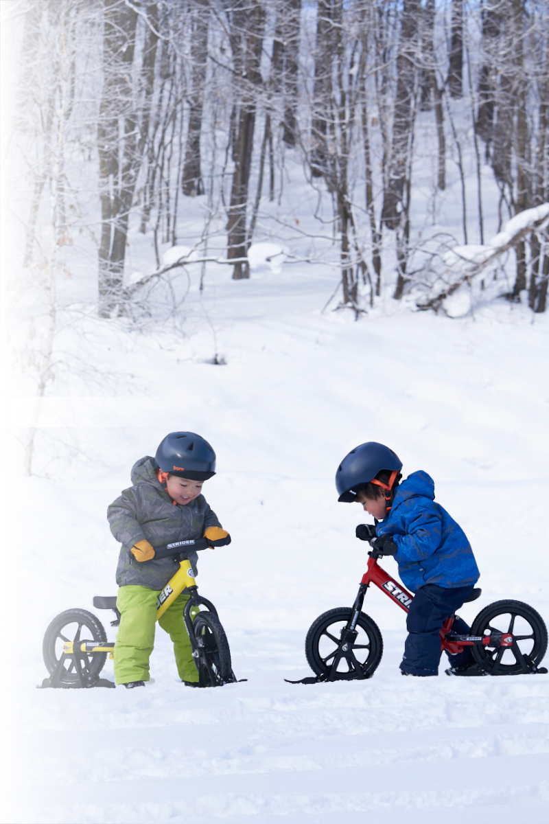 Two young riders wearing snowsuits, mittens and helmets straddle their Strider 12" Balance Bikes as they move through a snowy trail in a wooded area.