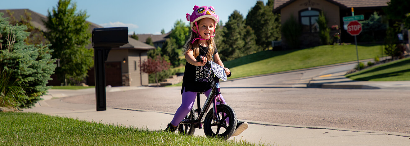 A child in a silly pink helmet rides a purple Strider 12 Pro balance bike down the sidewalk in a residential setting