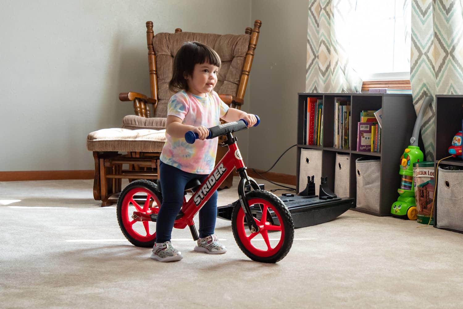 A child practices striding indoors on a red Strider 12 Sport balance bike with red Ultralight Wheels