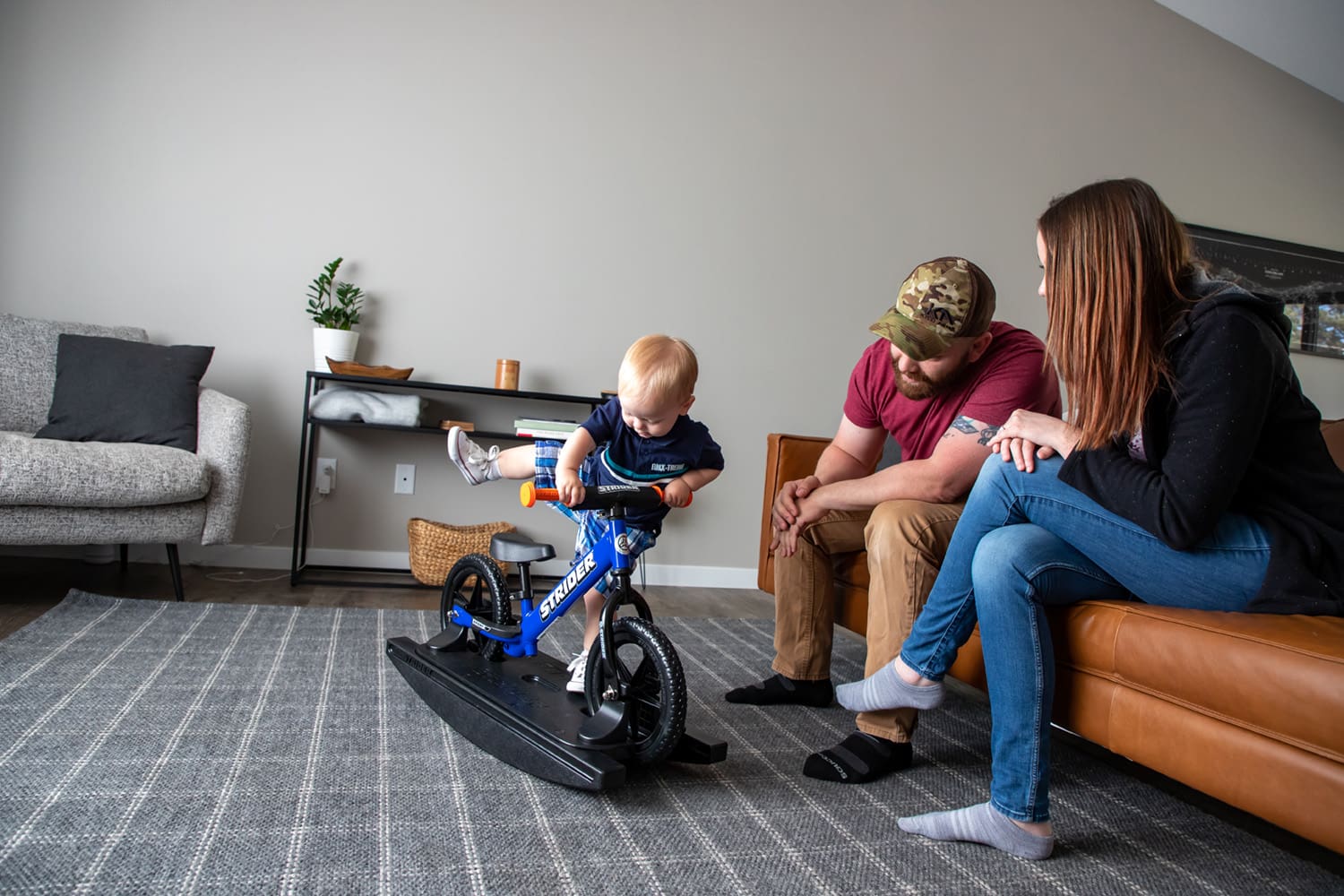 Parents watch from the couch while child plays on blue Strider Sport Rocking Bike