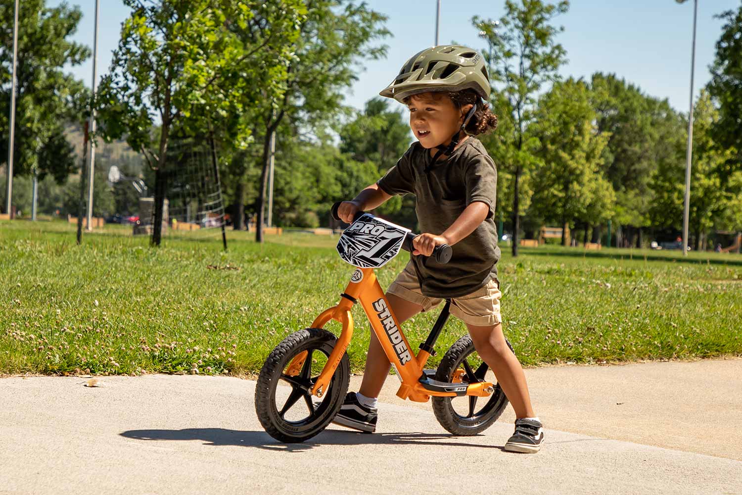 A by rides an Orange Rush Strider 12 Pro balance bike through a park