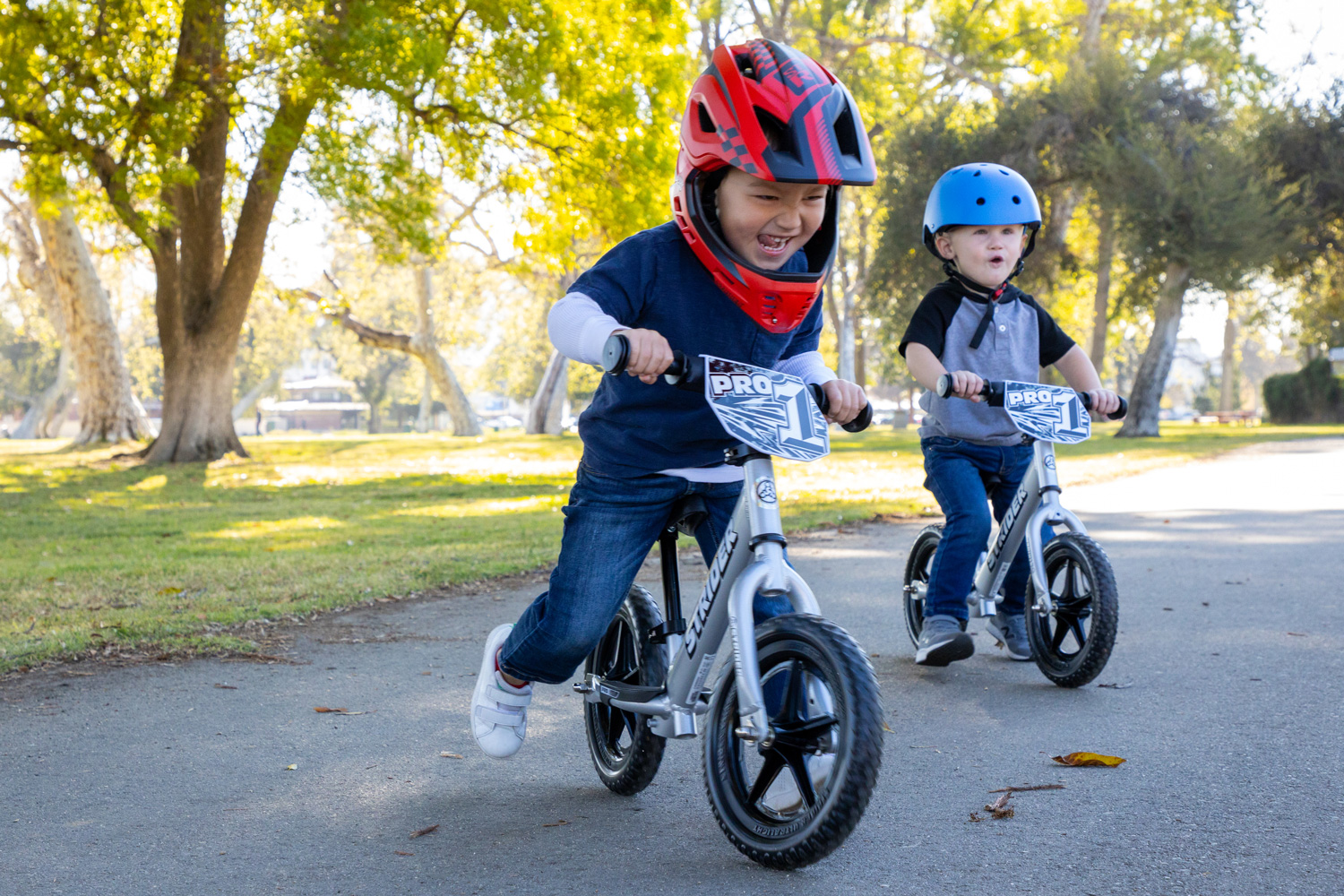 A boy in a red Strider ST-R bicycle helmet riding a silver Strider 12 Pro balance bike